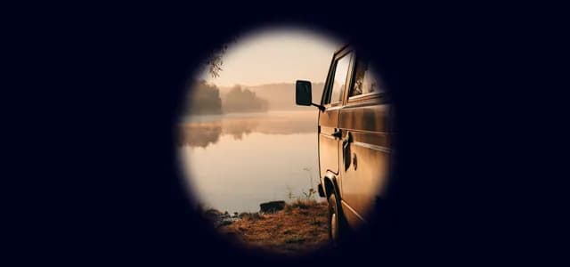 A parked van beside a calm lake at sunrise, viewed from the side with trees reflected in the water.