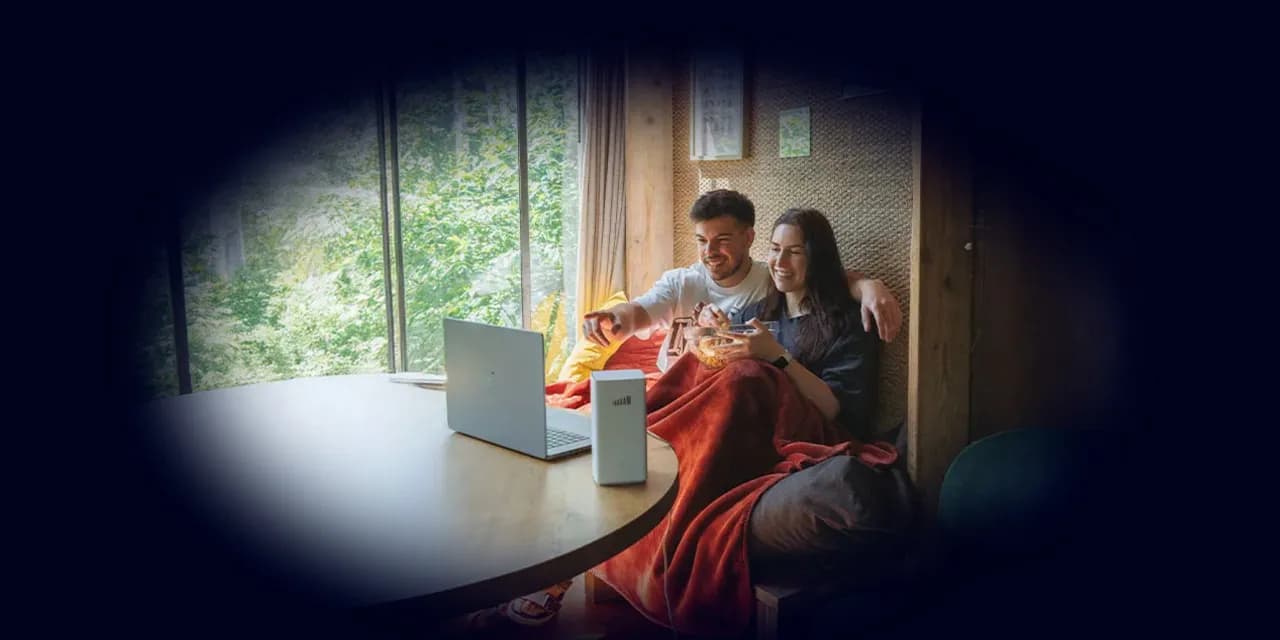 A smiling couple sits by a large window with trees outside, sharing snacks under a red blanket while watching a laptop together.