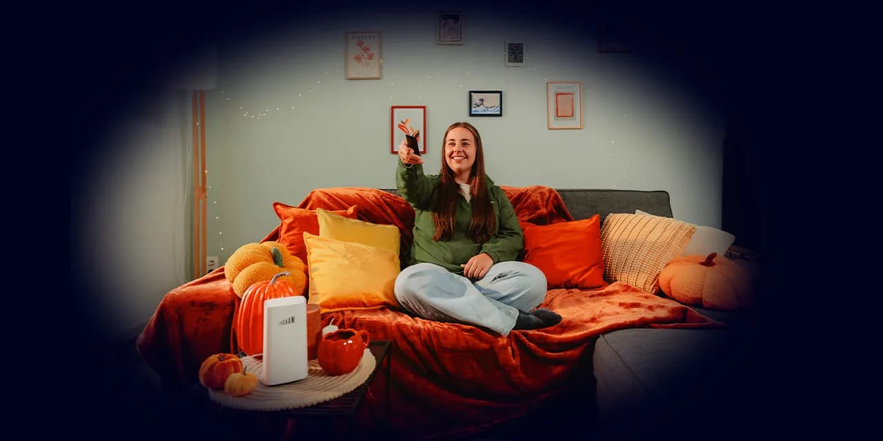 Smiling lady sitting on a cozy sofa with autumn-themed decorations, holding a remote control, surrounded by orange pillows, blankets, and pumpkin decor.
