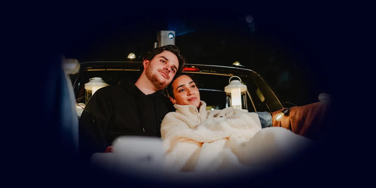 A couple sits wrapped in blankets in the back of a pickup truck at night, surrounded by string lights and lanterns. They appear to be watching a movie projected from a device mounted on the truck, creating a cozy outdoor cinema experience under a clear, starry sky.