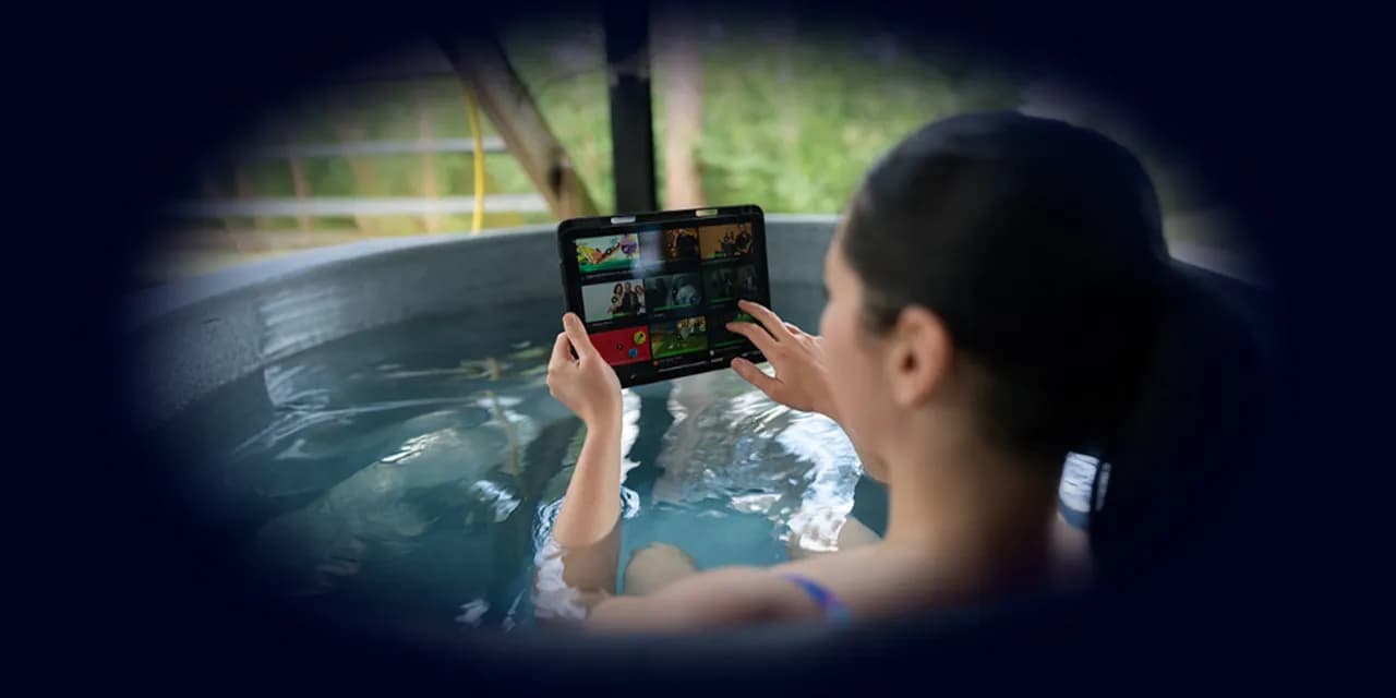 A person relaxes in a round outdoor hot tub, holding a tablet displaying streaming service thumbnails. The spa setting includes lush greenery and wooden structures, creating a serene wellness retreat atmosphere.