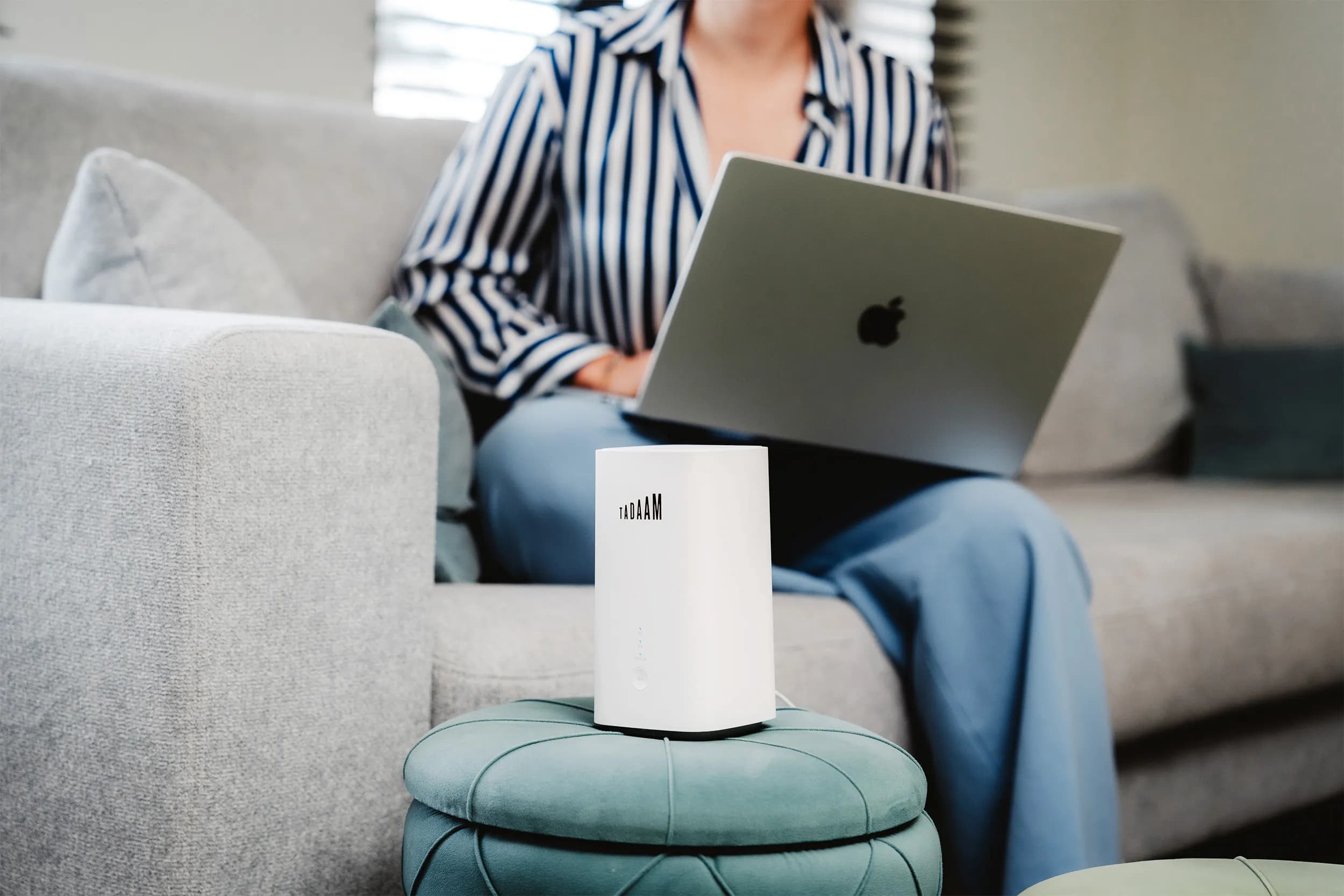 An image of a woman sitting on the couch with a Macbook laptop on her lap and a TADAAM's modem next to her.