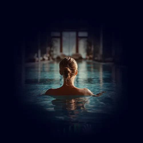 A person seen from behind, relaxing in a swimming pool inside a calm, softly lit space.