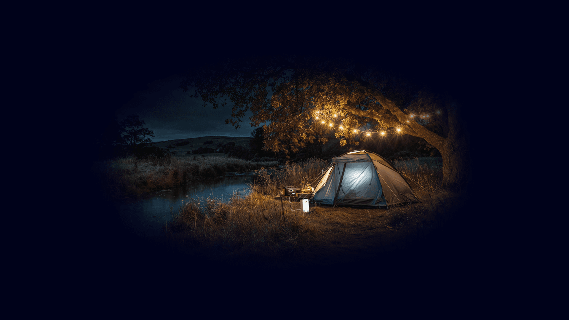 Tent glowing at night by a river, with string lights in a tree and a TADAAM modem set up outside — internet wherever you camp.