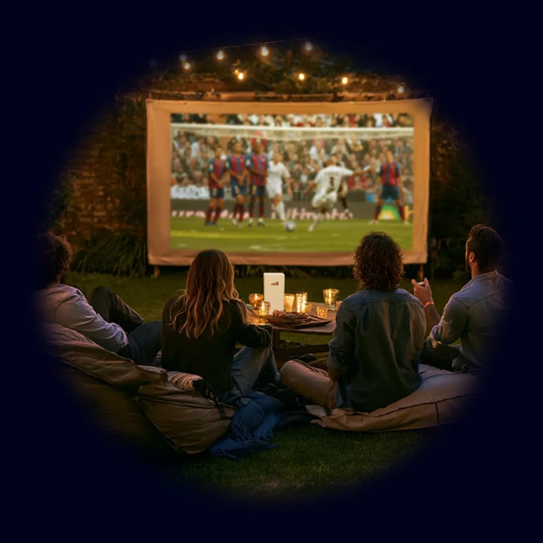 Friends watching a football match on an outdoor projector screen in a garden at night, with string lights and a TADAAM modem on the table.
