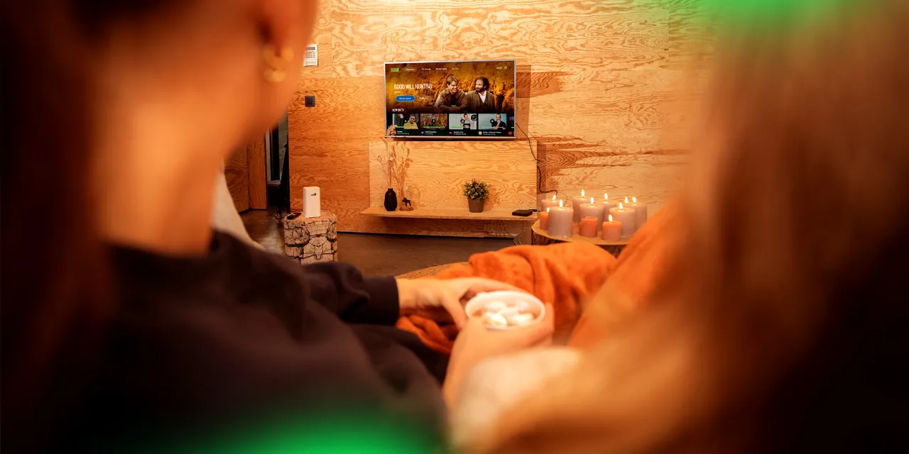 Two people relaxing on a couch with candles lit, watching Good Will Hunting on a TV in a cozy wooden room.