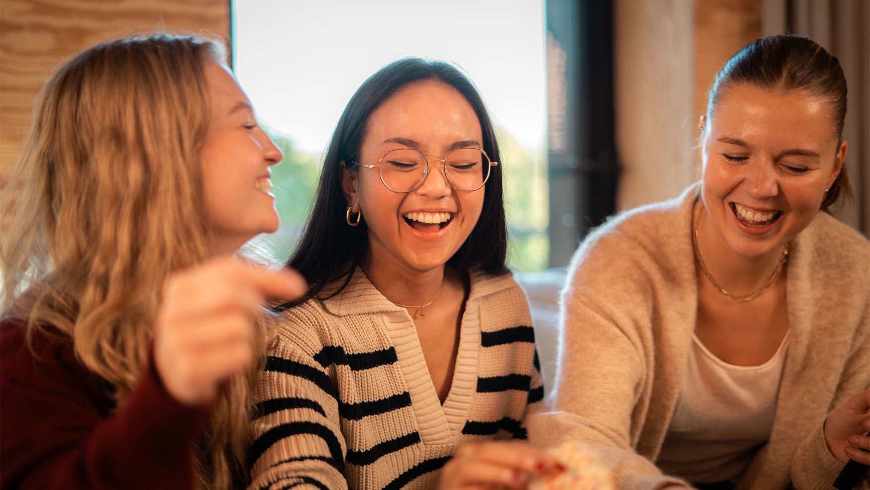 Three people sitting indoors, smiling and laughing together in a cozy, warmly lit setting.