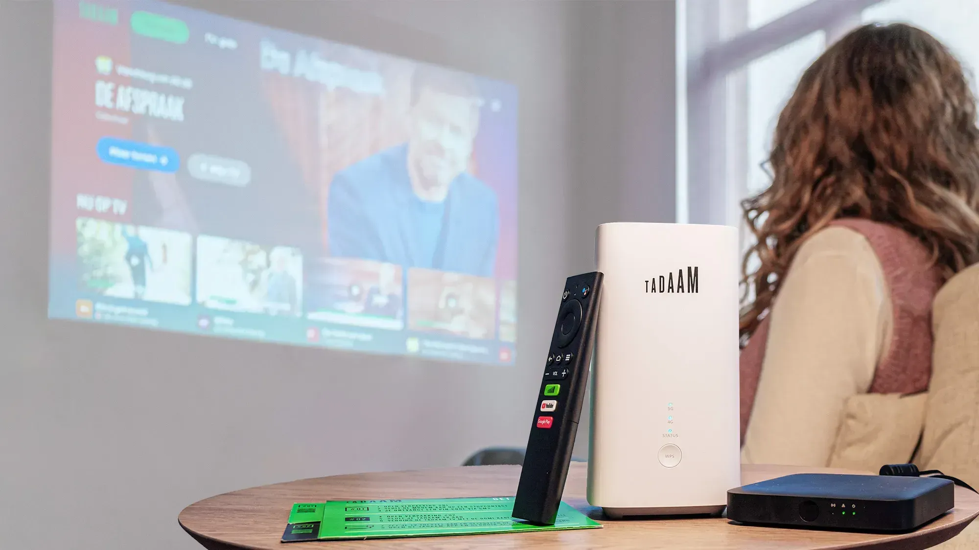 TADAAM's remote and a router placed on a table with streaming content projected on a wall, showing a cozy environment.